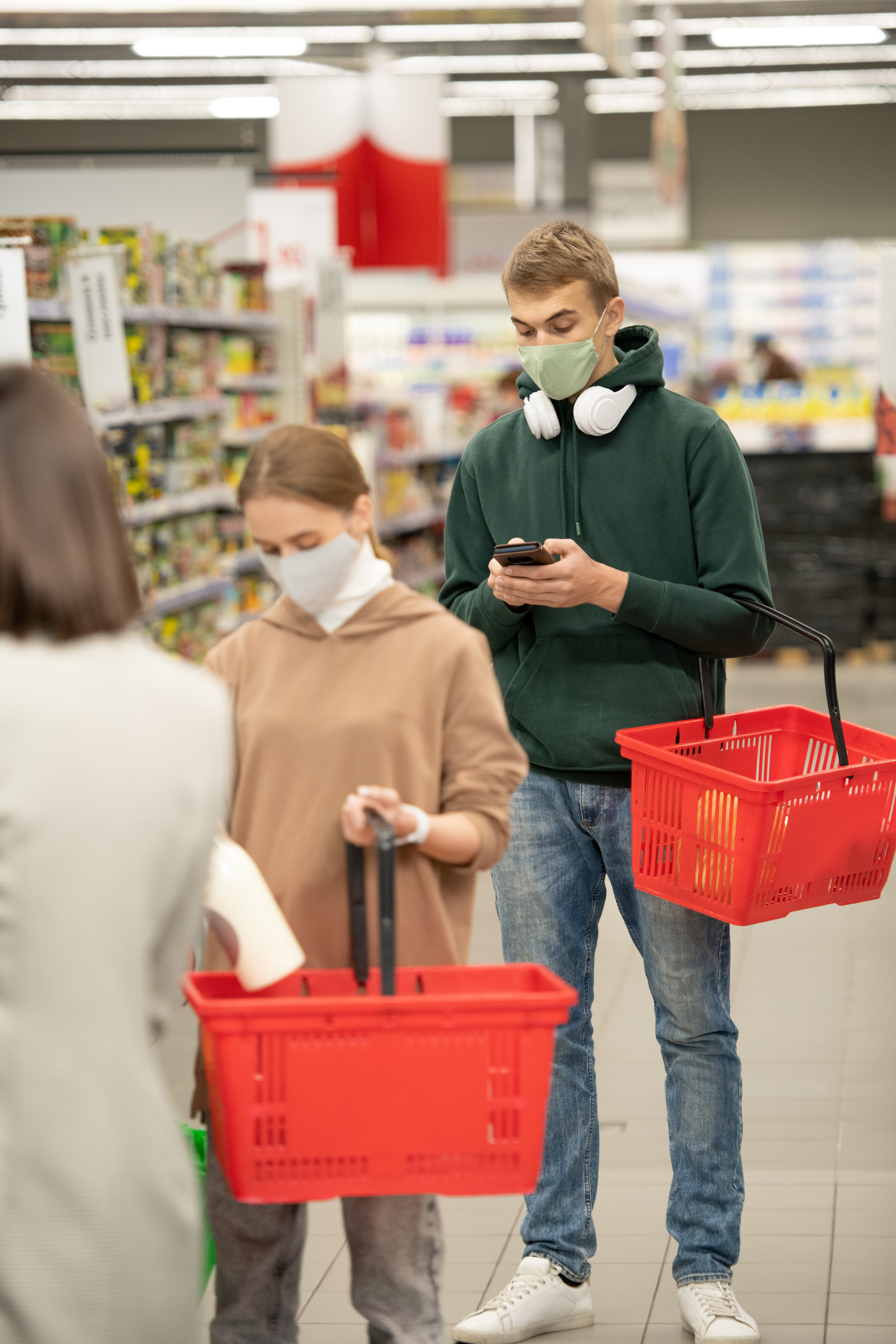 Several customers with plastic shopping baskets standing in queue by checkout counter in supermarket, one of them scrolling in smartphone